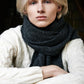 A close-up of a blonde man looking directly at the camera. He is wearing a cable-knit sweater, and his neck is bundled in a thick, fluffy dark grey wool scarf. The man is sitting with his arms crossed in front of him, and the lighting and clothing create a cozy, warm, and calm atmosphere.