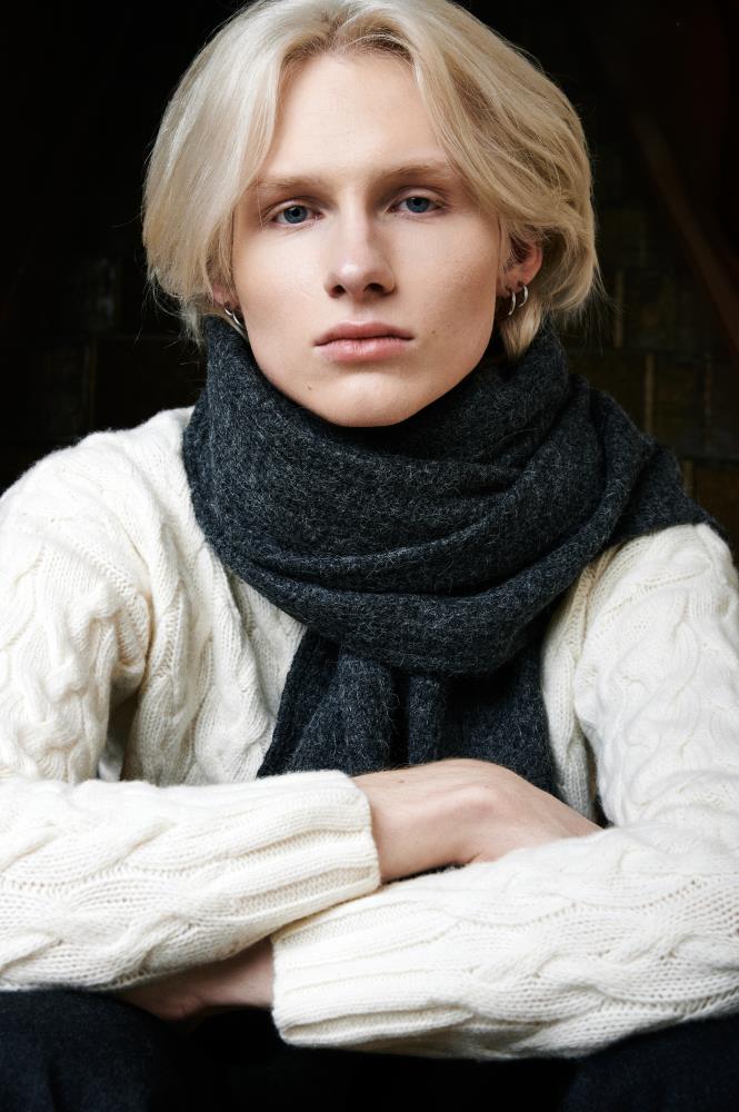 A close-up of a blonde man looking directly at the camera. He is wearing a cable-knit sweater, and his neck is bundled in a thick, fluffy dark grey wool scarf. The man is sitting with his arms crossed in front of him, and the lighting and clothing create a cozy, warm, and calm atmosphere.