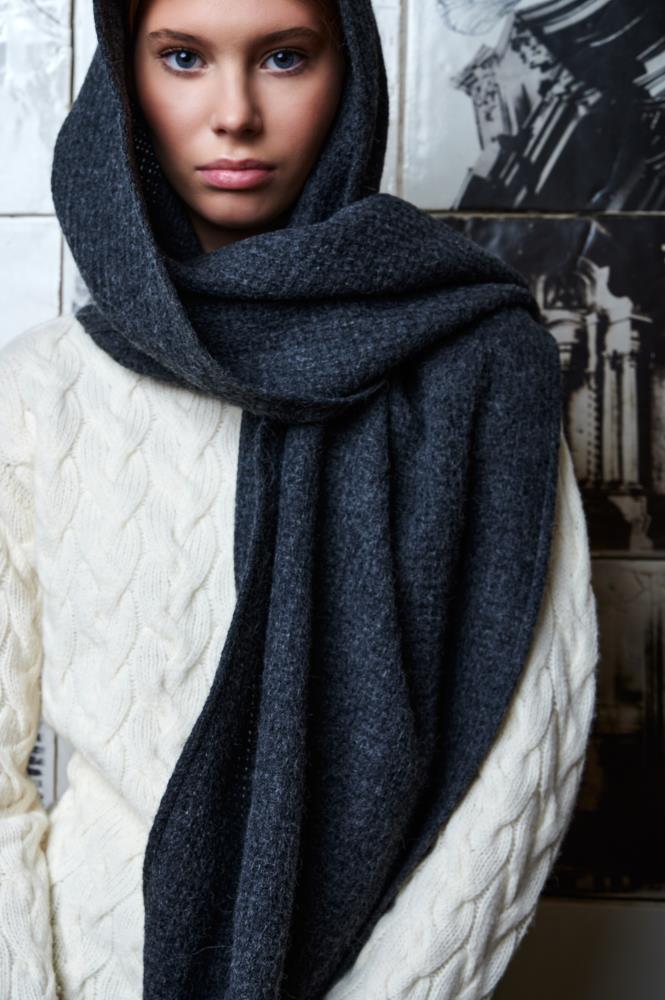A close-up of a young woman wearing a light cable-knit sweater. Her head and neck are wrapped in a thick, dark grey wool scarf, draped over her head like a hood with the end falling freely in front. The woman's gaze is steady and calm, and the background features textured tiles with architectural motifs, creating an artistic and cozy atmosphere.