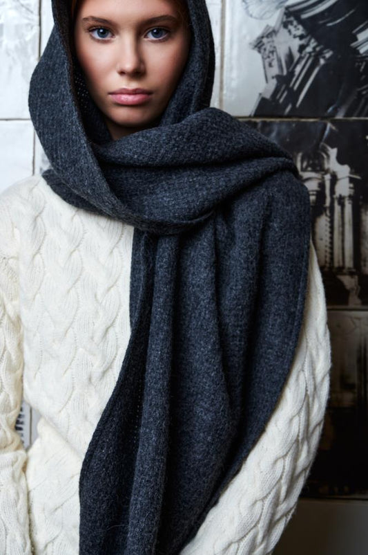 A close-up of a young woman wearing a light cable-knit sweater. Her head and neck are wrapped in a thick, dark grey wool scarf, draped over her head like a hood with the end falling freely in front. The woman's gaze is steady and calm, and the background features textured tiles with architectural motifs, creating an artistic and cozy atmosphere.