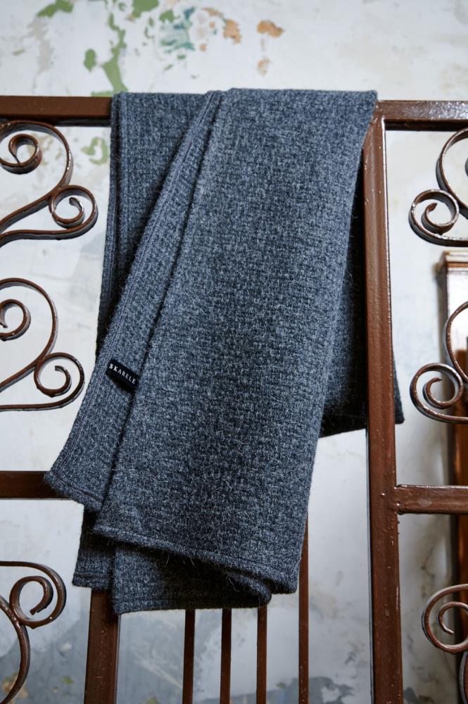 A dark grey wool scarf draped over brown metal railings with ornamental swirls. A small black label with the word "SKARELE" is attached to the edge. The background features a textured, distressed light wall with hints of blue and green, giving the image an industrial, vintage style.