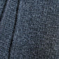 An extreme close-up of a dark grey wool fabric texture. The photo clearly highlights the dense knit or weave and the natural fluffiness of the material, conveying a sense of softness and coziness. A neat seam and a portion of a black label are visible on the left side, emphasizing the quality and detail of the accessory.
