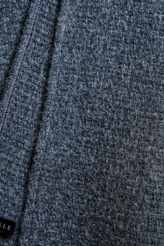 An extreme close-up of a dark grey wool fabric texture. The photo clearly highlights the dense knit or weave and the natural fluffiness of the material, conveying a sense of softness and coziness. A neat seam and a portion of a black label are visible on the left side, emphasizing the quality and detail of the accessory.
