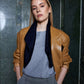 Woman with straight blonde hair stands confidently, wearing a tan eco leather jacket over a gray top and patterned pants, and navy silk scarf drapes loosely around her shoulders . She has hands in pockets, against a green marble wall.