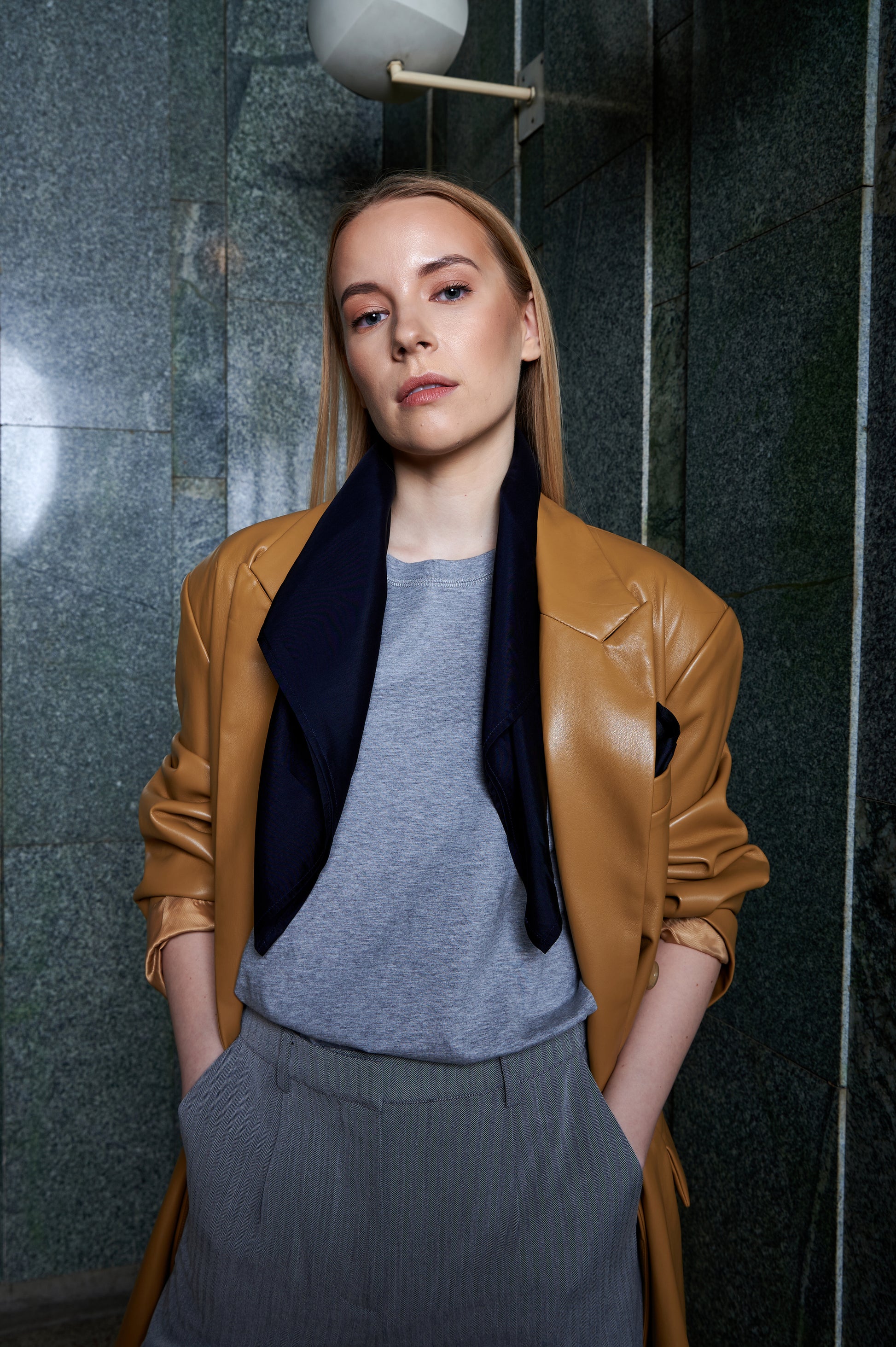 Woman with straight blonde hair stands confidently, wearing a tan eco leather jacket over a gray top and patterned pants, and navy silk scarf drapes loosely around her shoulders . She has hands in pockets, against a green marble wall.