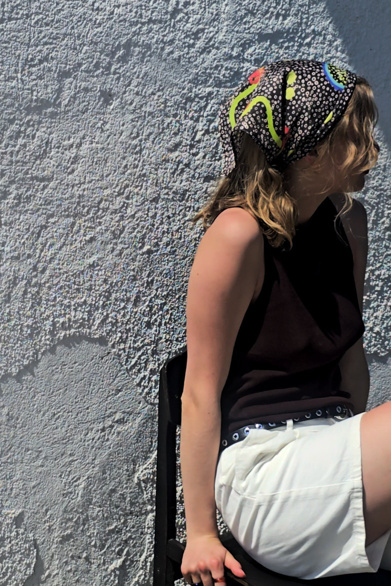 Woman styling the dark greyish-brown La Dolce Vita silk bandana on her head. Summer lifestyle fashion shot showcasing a luxury patterned headscarf on a sunny day.