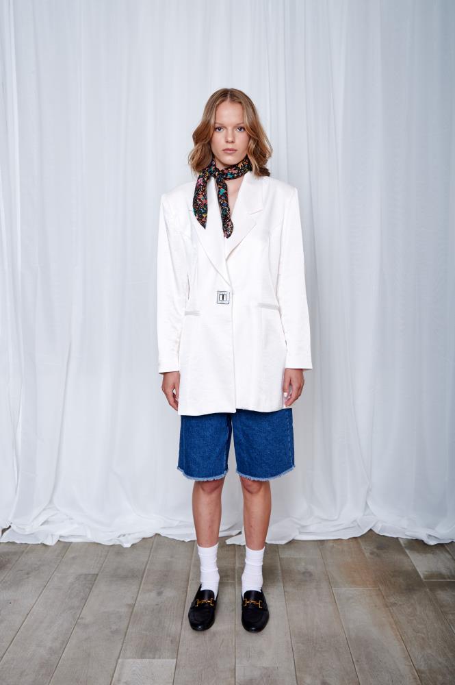 A woman in a white blazer and blue shorts stands on a wooden floor. She wears a colorful scarf, black loafers, and white socks, with a neutral expression.
