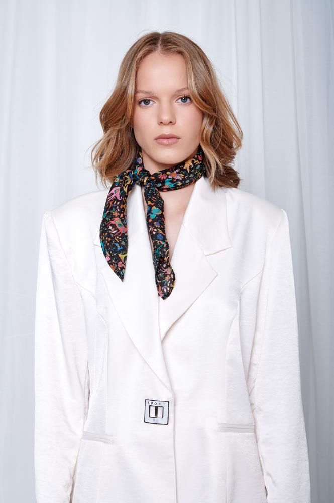 A woman with wavy blonde hair wears a white blazer and a colorful scarf. She poses against a light background, conveying a confident, poised demeanor.