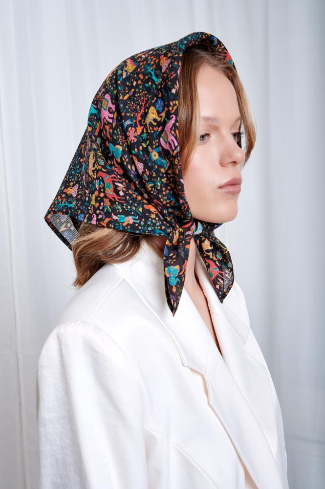 A person in a white blazer wears a colorful, patterned headscarf with floral motifs. The neutral background highlights a calm, stylish expression.