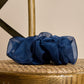 A voluminous, dark blue sheer fabric scrunchie placed on the woven seat of a wooden chair. The light and abundant fabric folds give the accessory a luxurious, airy look. The background is warm and neutral, and the texture of the wooden chair creates a cozy atmosphere.