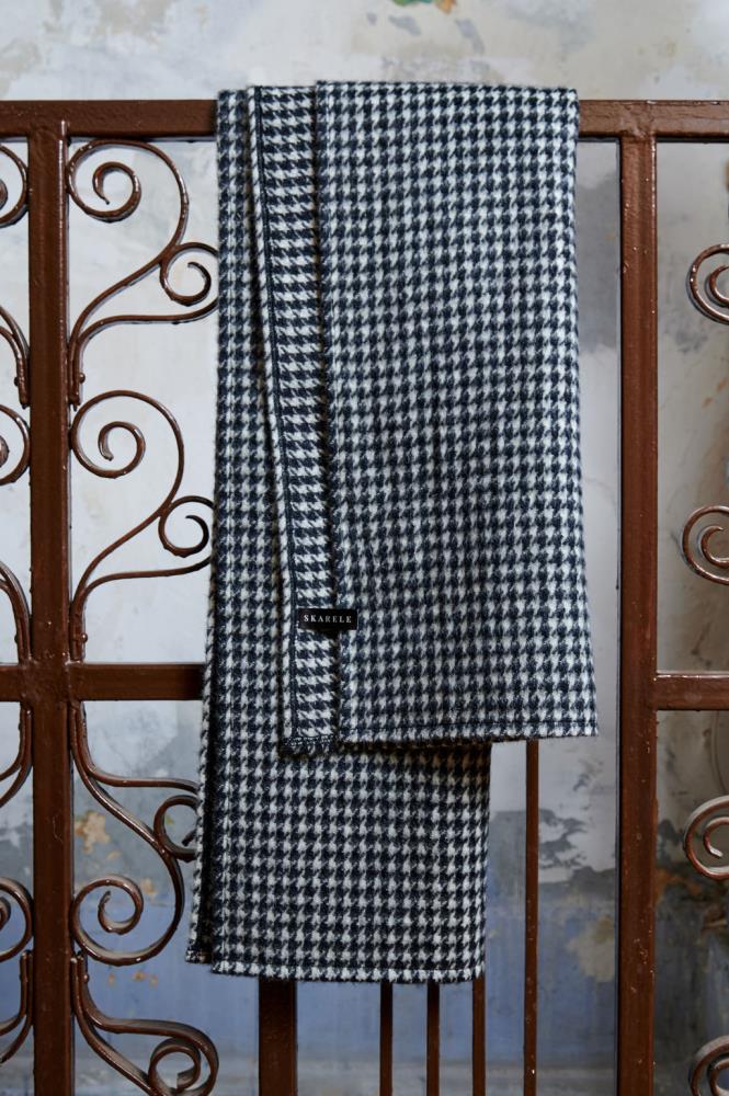 A wool scarf with a classic black and white houndstooth pattern draped over brown metal railings with ornamental swirls. A small black label with the word "SKARELE" is visible on the scarf. The background features a textured, light blue and grey wall, giving the image an industrial and slightly vintage feel.