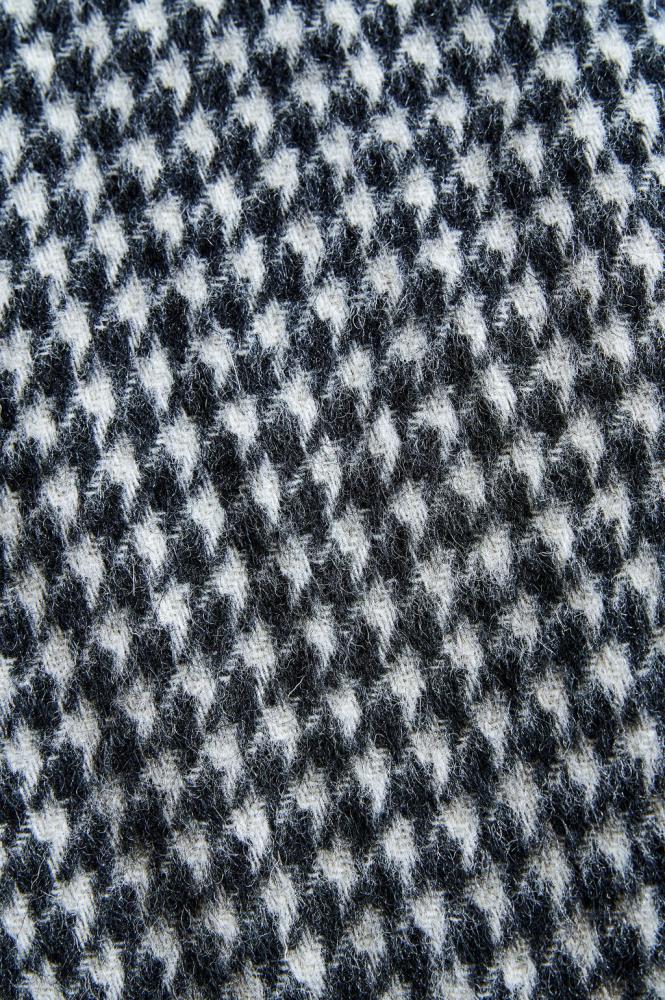an extreme close-up of a wool fabric texture featuring a classic houndstooth pattern. The photo clearly shows the dense weave and the natural fuzziness of the material. The contrast between black and white creates a dynamic, graphic look that emphasizes the high quality of the product and the coziness of the fabric.