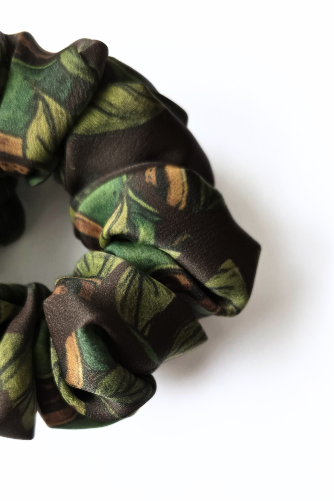  A detailed close-up of a luxury patterned silk scrunchie, showcasing vibrant green leaf motifs and rich brown details on the fabric folds