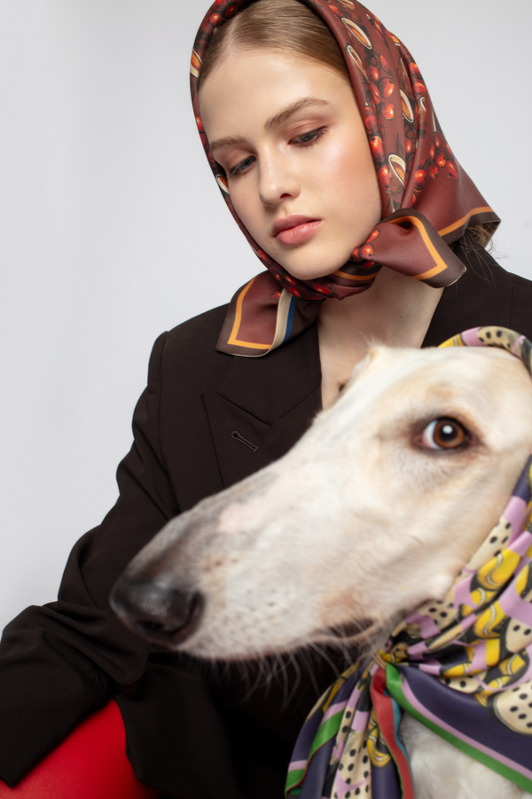 A portrait featuring a woman in a brown blazer wearing a coffee-themed silk headscarf. Beside her is a Borzoi dog wearing a vibrant, colorful patterned scarf. White background.