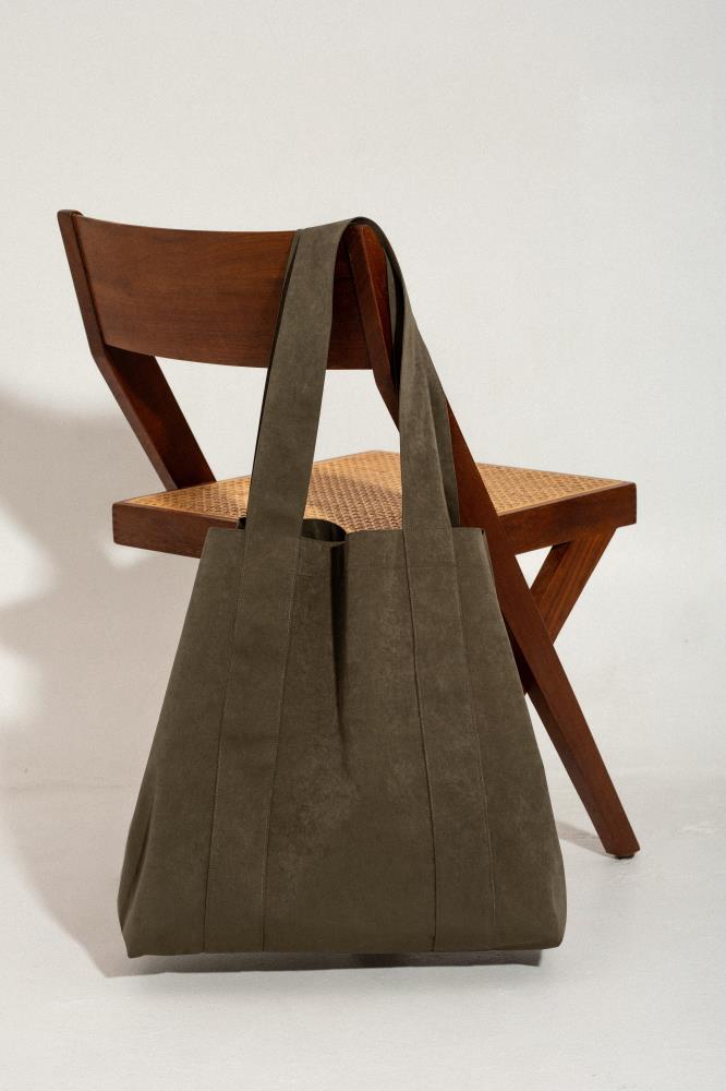 A front view of an oversized dark olive green canvas tote bag with long handles, positioned next to a vintage wooden chair with a rattan seat. Set against a white studio background.