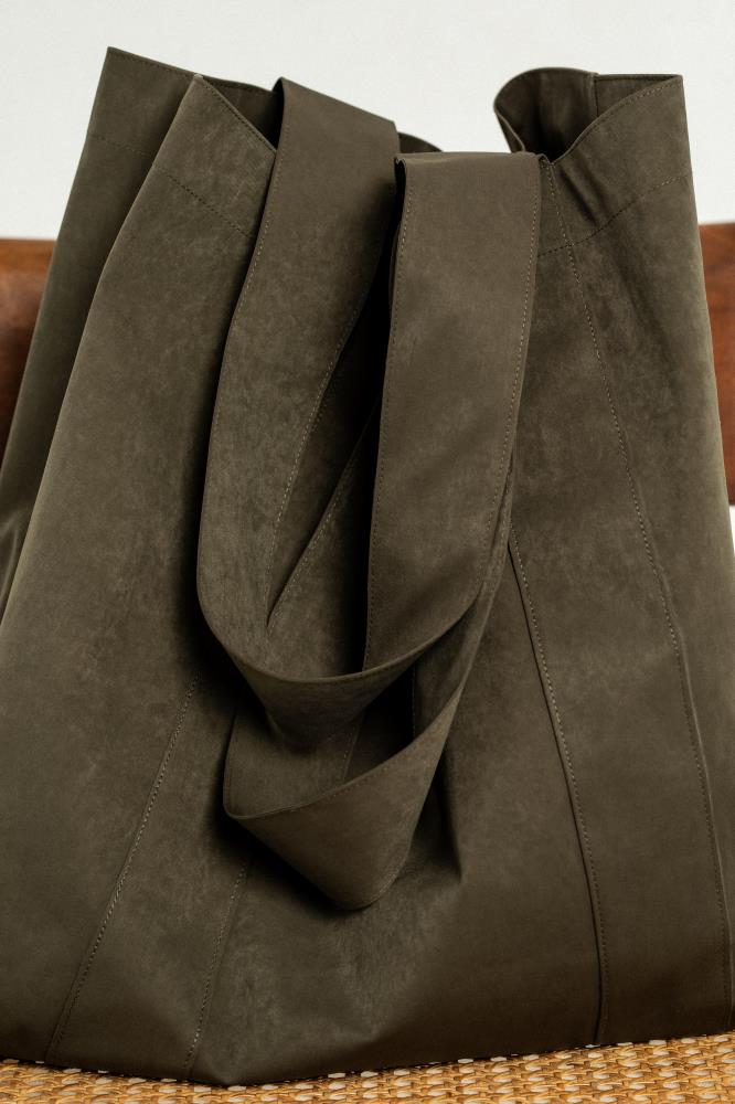 A close-up view of a dark olive green, textured oversized canvas tote bag, showcasing wide matching shoulder straps, subtle vertical stitching, and a portion of a rattan-seat chair below.