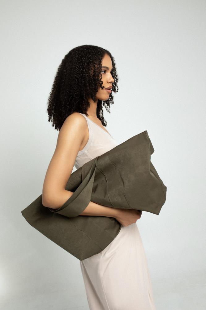 A profile view of a woman model with long curly hair, wearing a neutral beige dress and holding a large, dark olive green oversized canvas tote bag against her chest. Set against a white background.