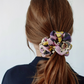 A rearview medium close-up shot captures a person with auburn hair in a messy ponytail, styled with a patterned silk scrunchie. The scrunchie features abstract prints of pink, yellow, purple, black, and polka dots. The individual is wearing a dark navy blazer and the background is a simple, light white wall.