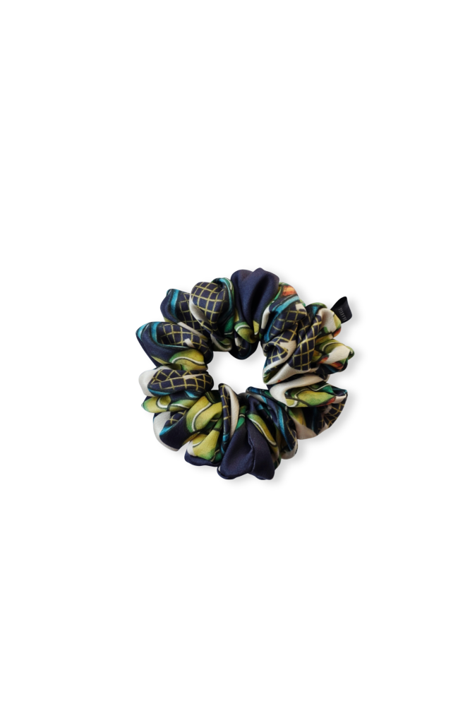 A top-down view of a luxury patterned silk scrunchie, displayed flat on a white background. It features a dark blue print with vibrant green, yellow, and red details and a small brand tag.