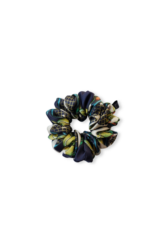 A top-down view of a luxury patterned silk scrunchie, displayed flat on a white background. It features a dark blue print with vibrant green, yellow, and red details and a small brand tag.