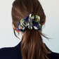 A rear view portrait of a woman model with her brown hair tied back in a ponytail, adorned with a luxury patterned silk scrunchie in blue, green, and yellow on a white background.