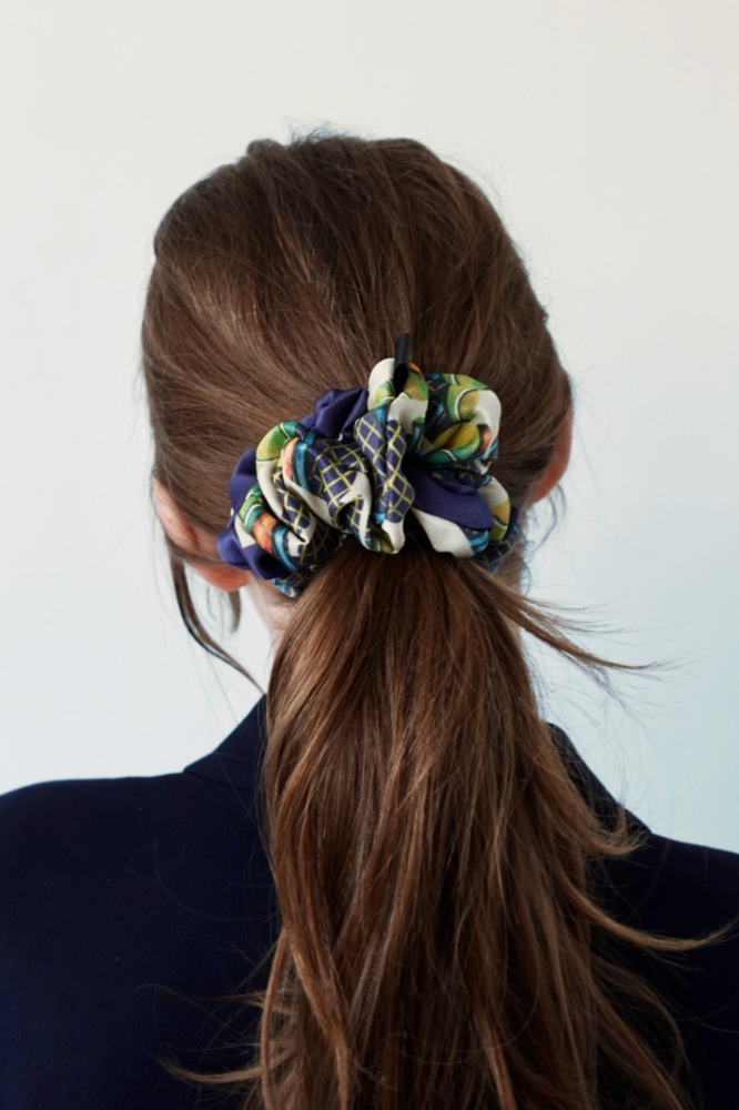 A rear view portrait of a woman model with her brown hair tied back in a ponytail, adorned with a luxury patterned silk scrunchie in blue, green, and yellow on a white background.