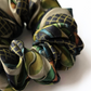 A detailed close-up of a luxury silk scrunchie, showcasing a sophisticated blue and green geometric pattern on twisted fabric folds. Displayed against a clean white background.