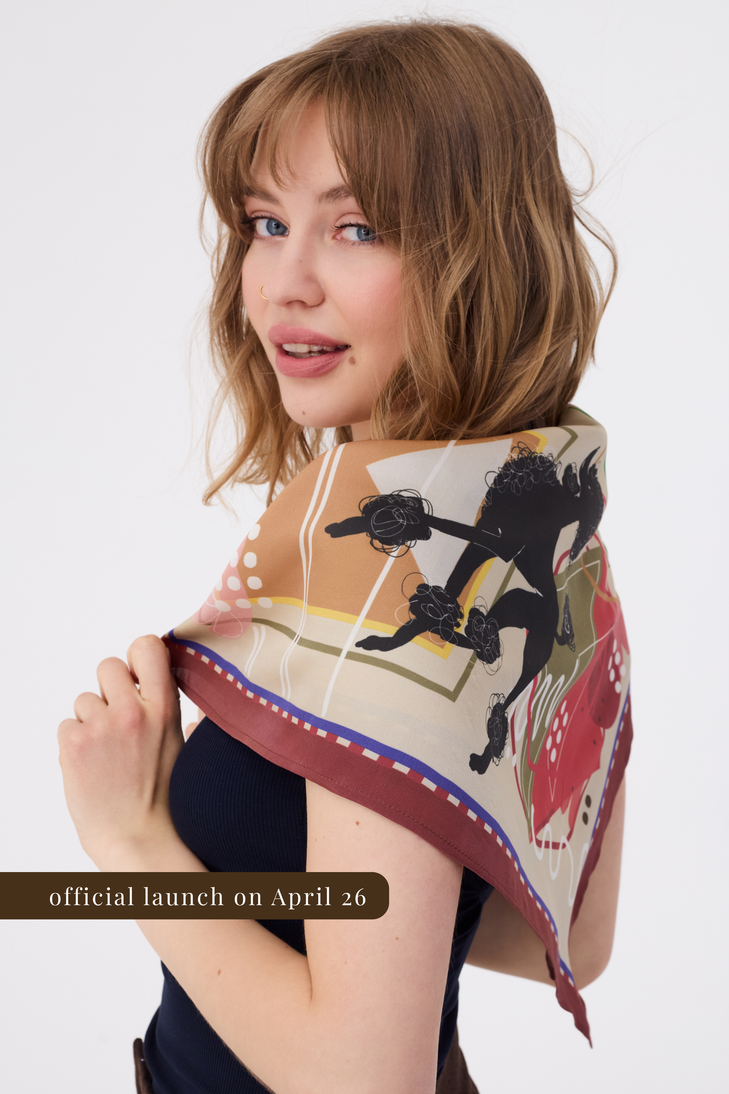 portrait of a woman looking over her shoulder while showcasing a silk scarf draped around her upper arms and chest. She is wearing a dark ribbed top against a white background. The scarf features an abstract illustration in warm, earthy tones with geometric elements and stylized animal motifs. A brown banner at the bottom left reads "official launch on April 26".