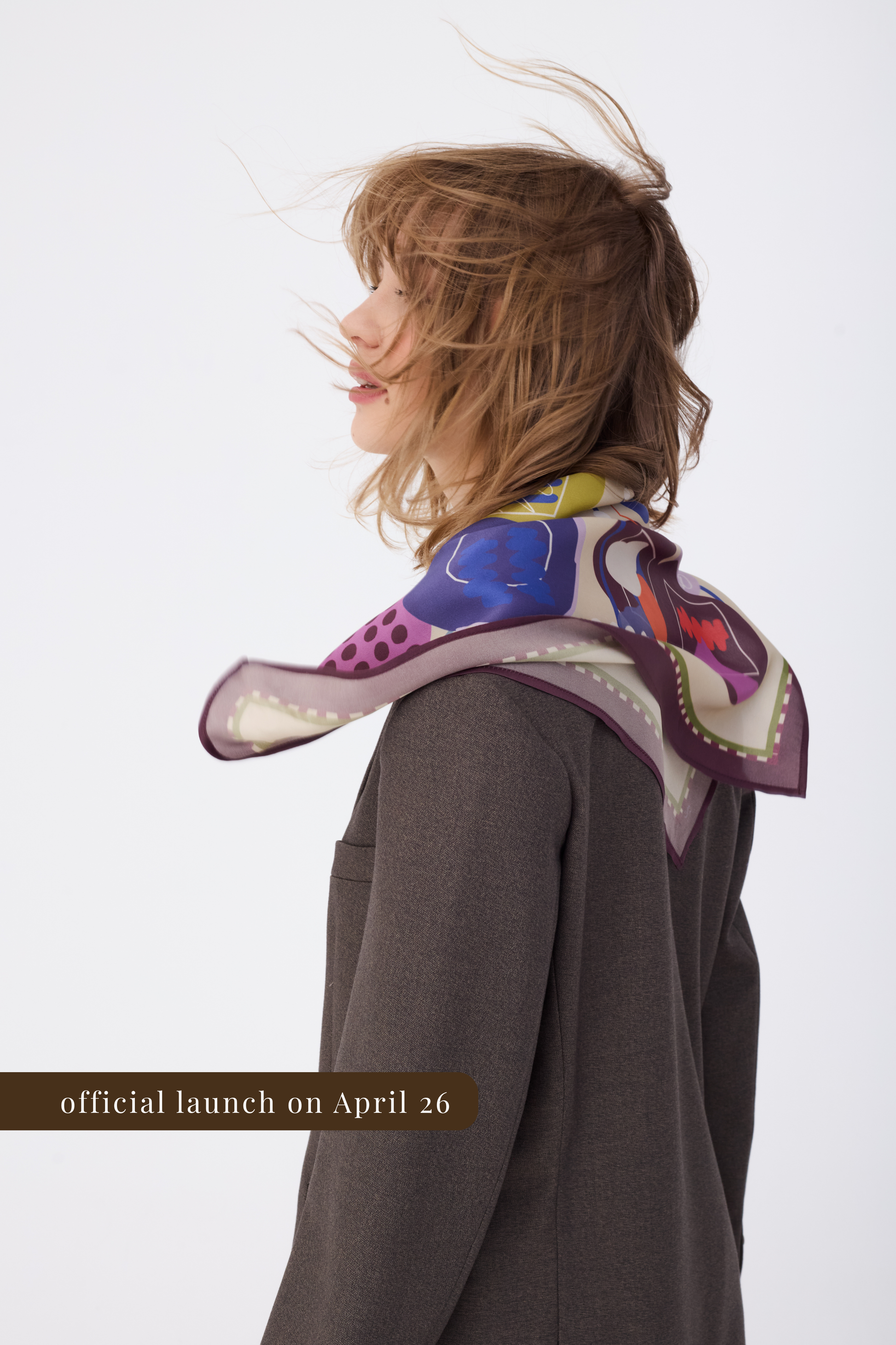 A profile view of a model wearing a textured dark brown blazer. A silk scarf with an abstract artistic pattern featuring blue, burgundy, olive, and purple shapes is draped over her shoulders.Patterned silk scarf with a plum border worn over a brown blazer, viewed in profile. The image focuses on the scarf's abstract design and drape. A text overlay indicates an April 26 launch date.