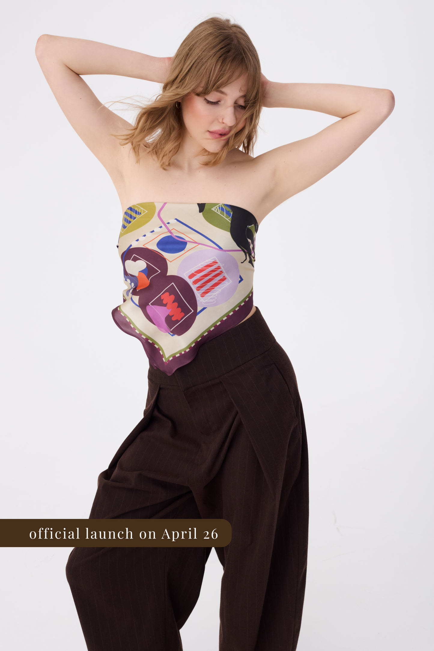 A woman wearing a large silk scarf styled as a strapless top, paired with high-waisted dark brown wide-leg trousers. The scarf features an abstract artistic print with purple, lilac, and orange geometric shapes on a cream base. A gold banner at the bottom reads "official launch on April 26".