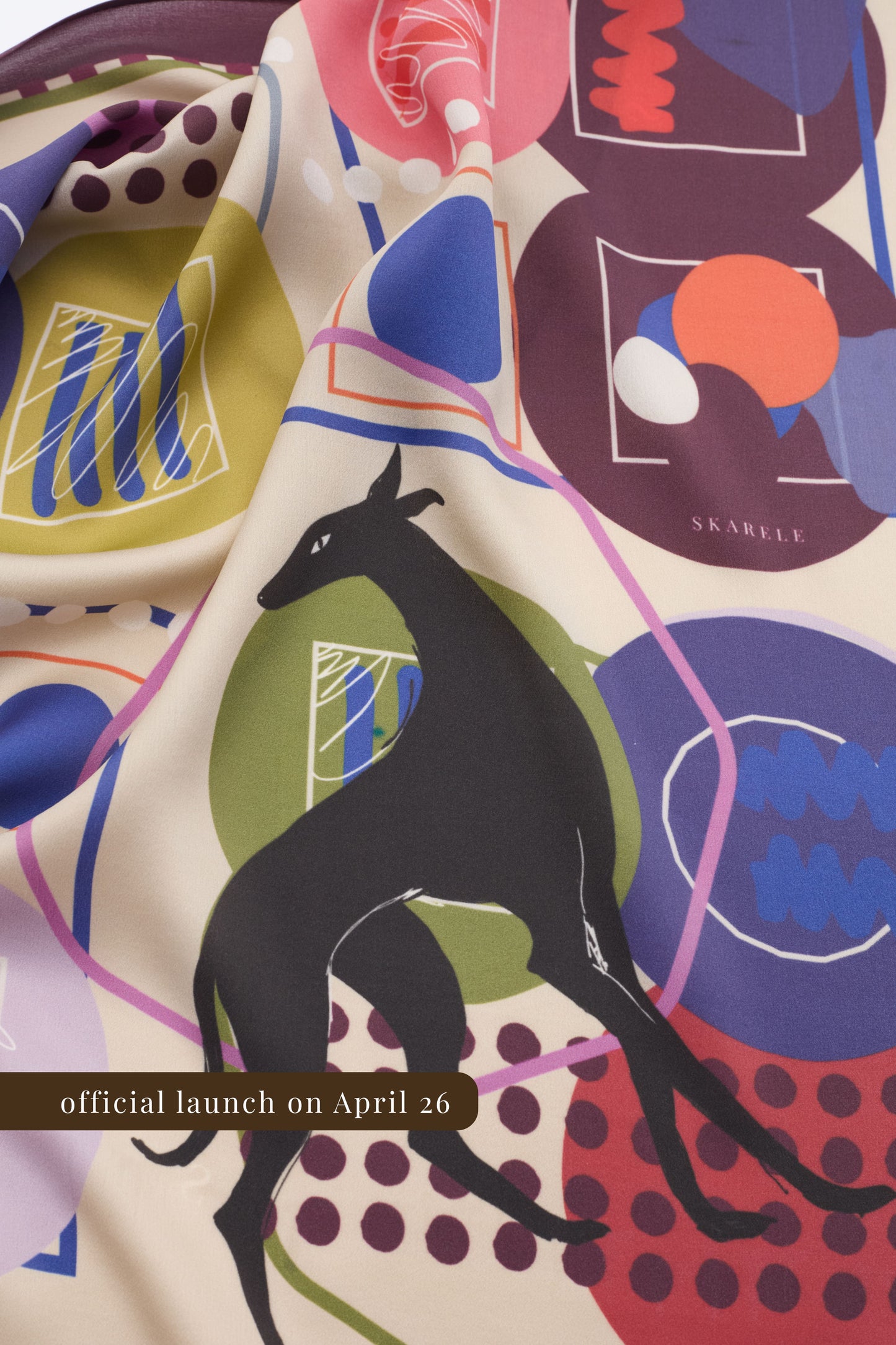 A close-up photograph of a draped silk scarf featuring an abstract artistic print with a central black greyhound illustration. The design includes colorful geometric shapes in pink, lilac, and olive green, with the brand name "SKARELE" subtly visible. A brown banner at the bottom indicates the official launch on April 26.
