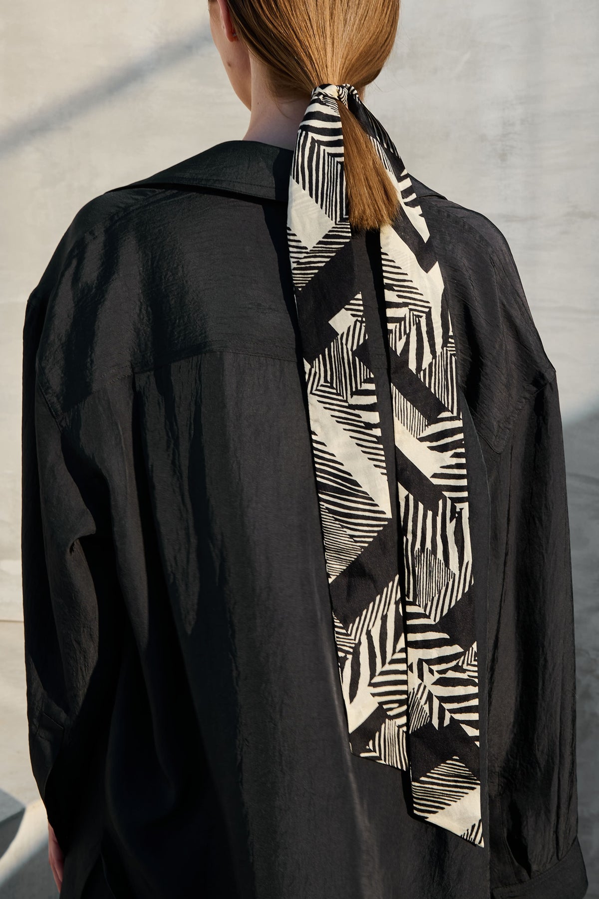 A back view photo focusing on a trendy narrow silk scarf stylishly tied into a woman's hair. The scarf features a graphic black and white botanical pattern and drapes contrastingly over a black shirt. This is a versatile hair accessory that adds a modern and elegant touch to the look.