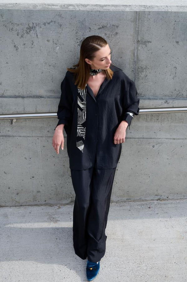 A model poses by a concrete wall, wearing a loose black suit and a narrow "Twilly" silk scarf tied around her neck. The scarf features a graphic black and white pattern, adding a modern accent to the minimalist look. Stylish blue velvet shoes complete the elegant and industrial aesthetic.