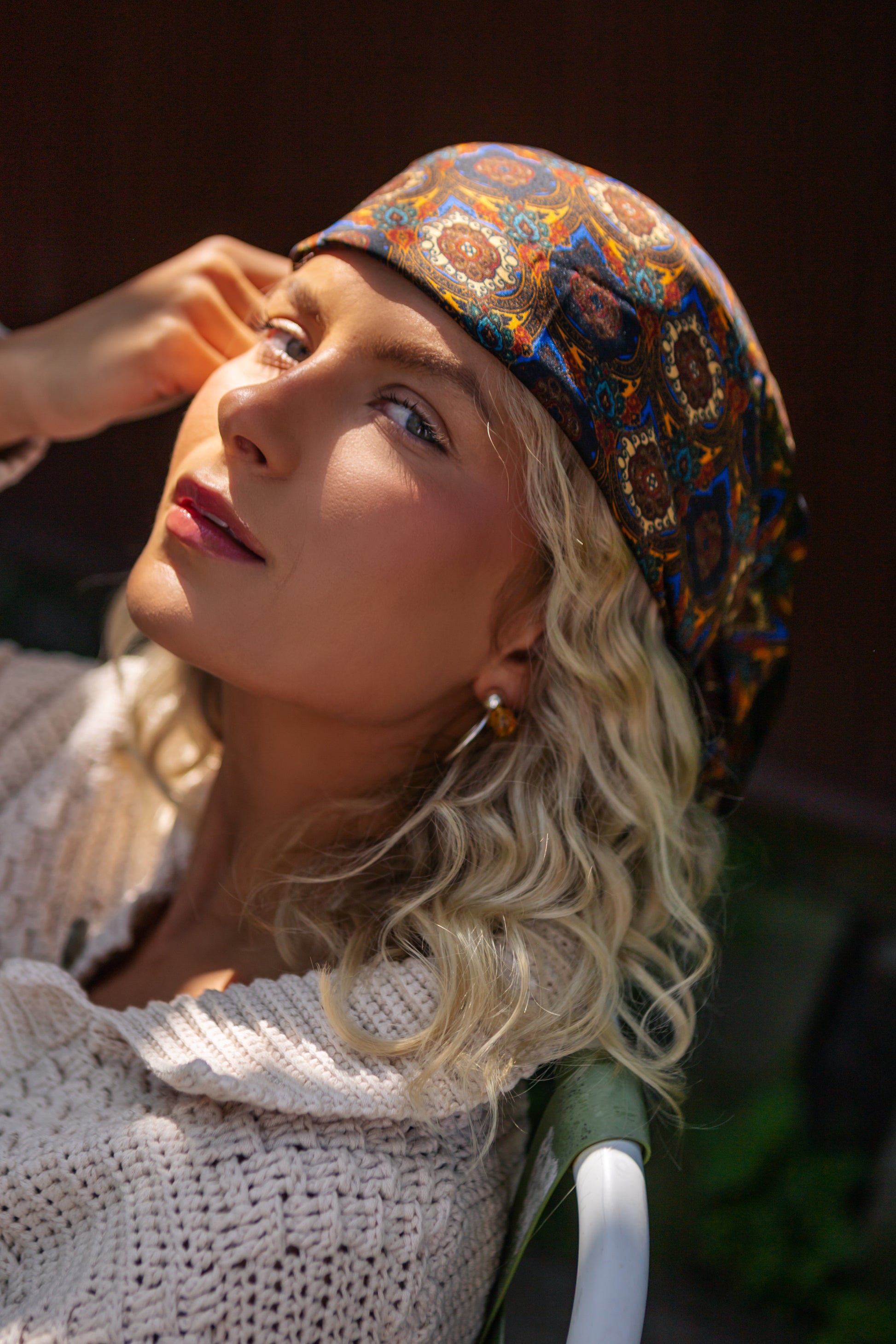radiant blonde woman basks in the sun, wearing a vibrant patterned bohemian silk headscarf and a cozy crocheted top, evoking a carefree, summery vibe.