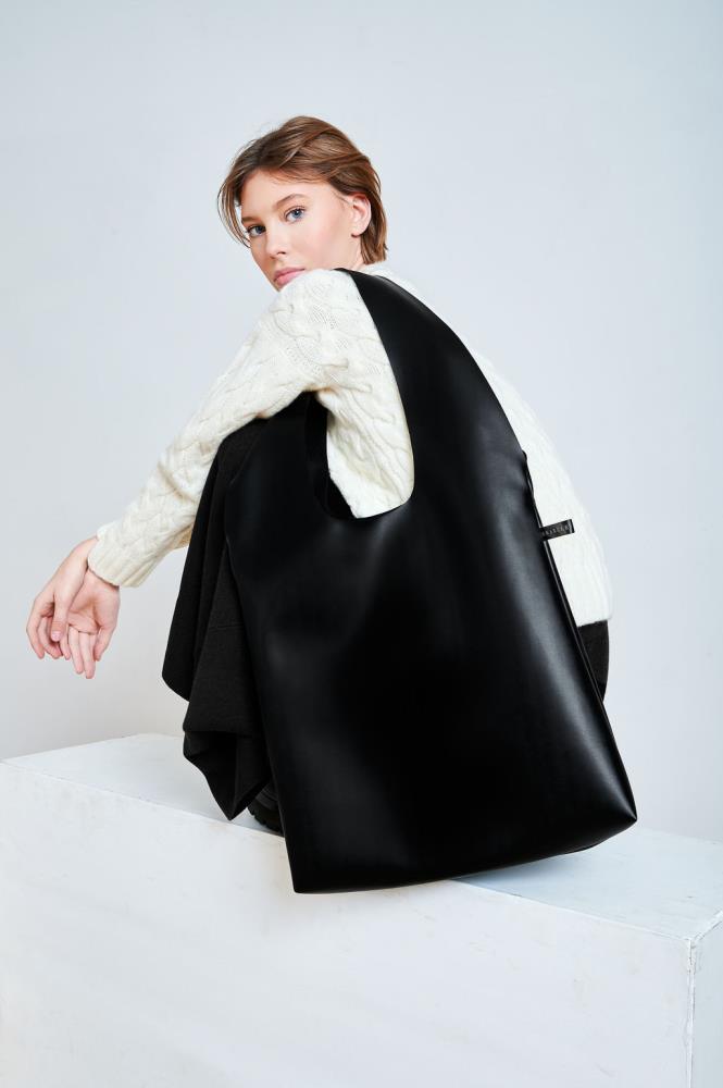 A large, slouchy black eco leather tote bag worn by a model in a white cable-knit sweater, showcasing its spacious design and minimalist urban aesthetic.