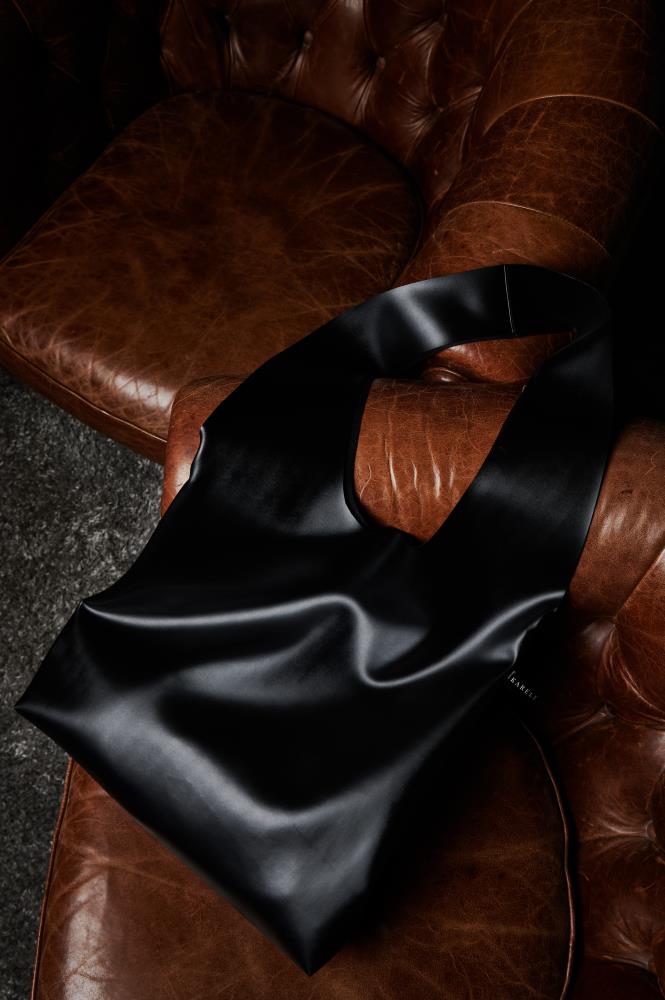 A sleek black eco leather tote bag draped over a vintage brown leather armchair, showcasing its smooth texture and elegant, minimalist silhouette in a rich, moody setting.