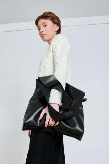 model posing with a large black eco leather tote bag, showcasing its oversized silhouette and smooth texture paired with a white cable-knit sweater for a modern urban look.