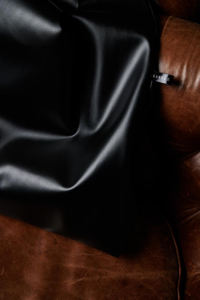A close-up of a premium black eco leather tote bag, focusing on the soft texture and elegant folds, featuring the "SKARELE" brand label against a rich brown leather armchair.