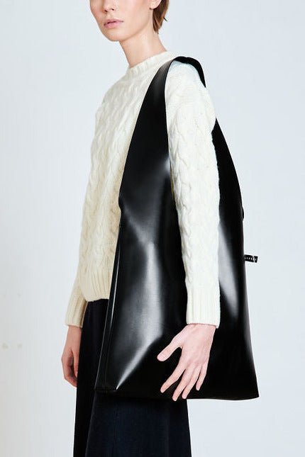 A side view of a large black eco leather tote bag worn by a model, showcasing its oversized, slouchy silhouette and premium smooth finish against a white cable-knit sweater.