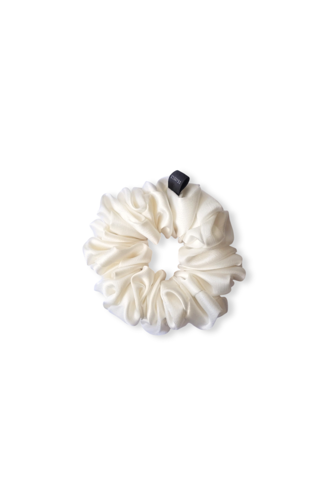 A top-down view of a luxury, white gathered silk scrunchie, displayed flat against a clean white background. It features a soft, lustrous texture and a small black tag.