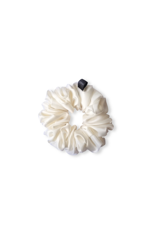 A top-down view of a luxury, white gathered silk scrunchie, displayed flat against a clean white background. It features a soft, lustrous texture and a small black tag.