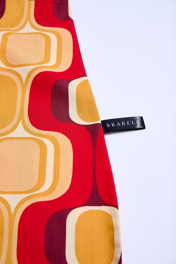 A close-up shot of a fabric item with a vibrant retro-style pattern against a clean white background. The design features wavy shapes and rounded squares in yellow, orange, red, and burgundy. A small black label with the white text "SKARELE" is visible on the right side.