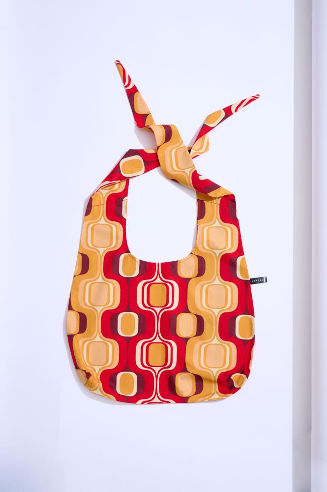 A vibrant textile tote bag photographed against a white background. The bag features a colorful retro-style pattern with rounded squares in yellow, orange, and red. Its handles are tied in a knot at the top, and a small black brand label is visible on the right side.