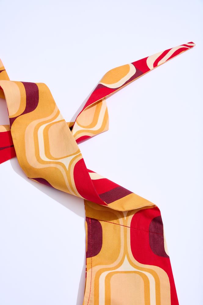 A close-up shot of a twisted and knotted fabric against a white background. The material features a vibrant retro-style pattern with rounded shapes in yellow, orange, red, and burgundy. The knot and folds create a dynamic texture and highlight the warm color palette.
