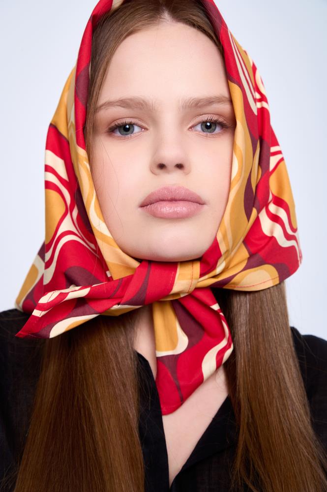 A young woman looking directly at the camera, wearing a vibrant cotton scarf over her head. The scarf features wavy retro patterns in red, yellow, and burgundy, tied in a knot under her chin. Her long, straight brown hair falls over her shoulders against a light, neutral background.