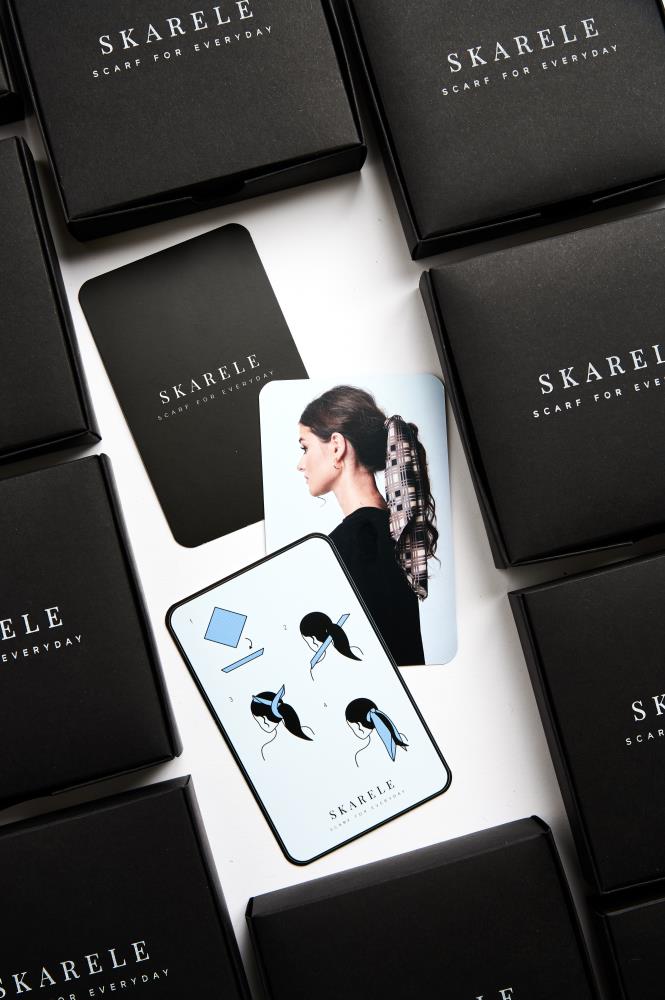 Close-up of SKARELE scarf tying cards featuring an illustrated, step-by-step guide on how to style scarves, surrounded by elegant black branded packaging.