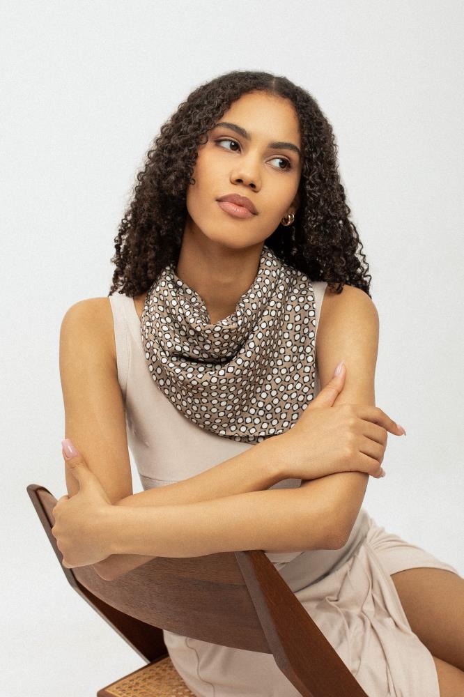 A portrait of a woman model with curly hair sitting on a wooden chair, wearing a viscose scarf with a brown and white geometric pattern bandana style. Set against a white background.

