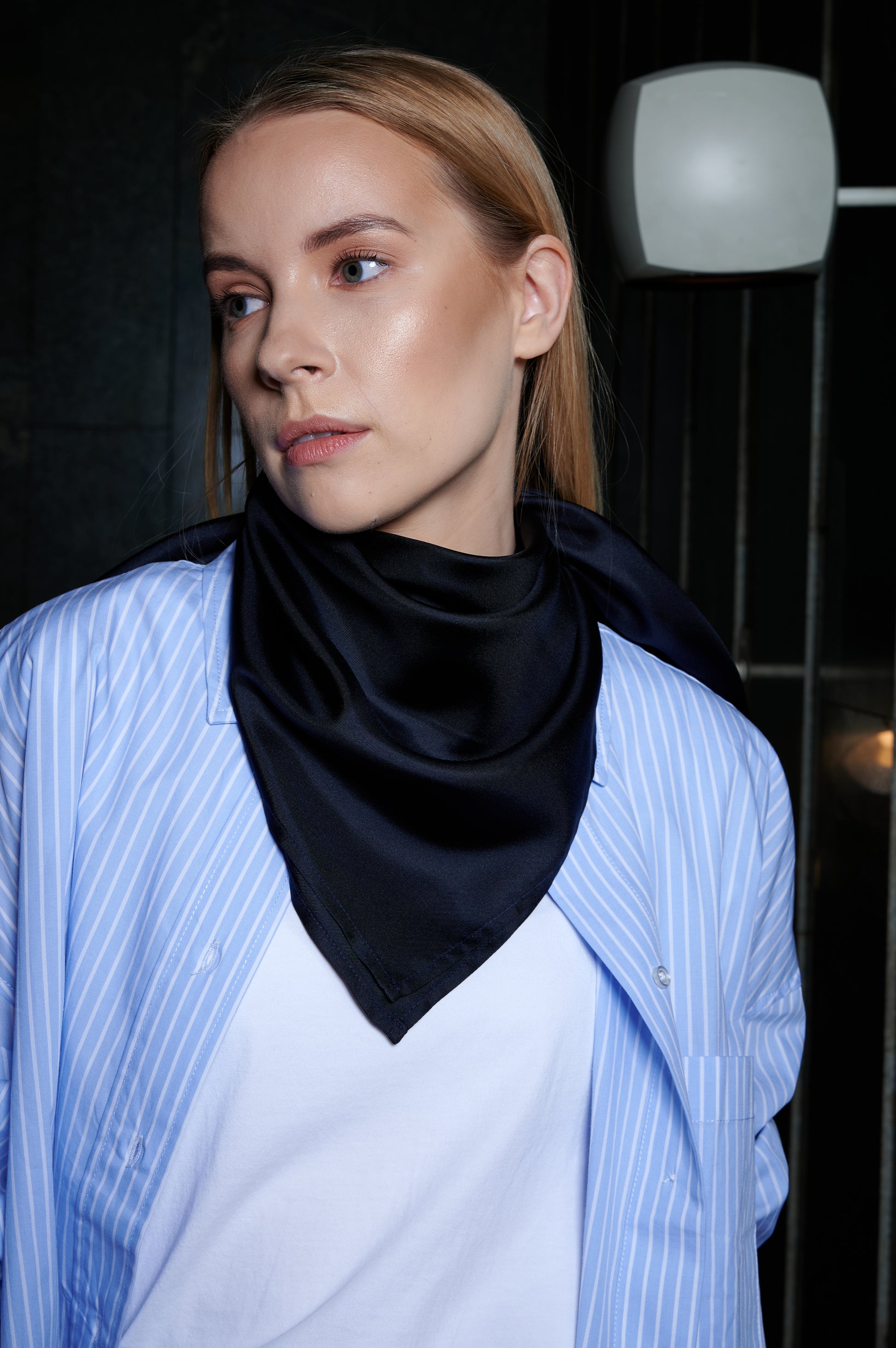 A woman with light hair gazes to the side, wearing a blue striped shirt and a midnight blue silk bandana style. The dimly lit background adds a contemplative mood.