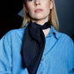 Portrait of a person with long blonde hair in a blue denim shirt, wearing a midnight blue silk scarf. The expression is calm and confident against a dark background