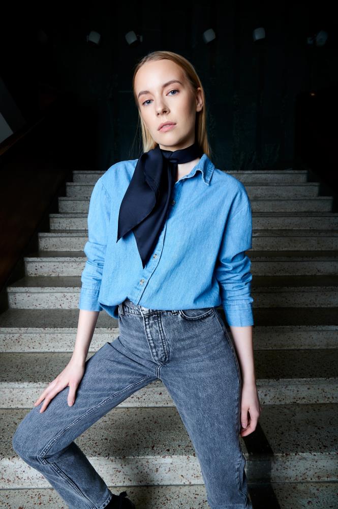 A person stands on stairs wearing a blue denim shirt, gray jeans, and a midnight blue silk scarf around the neck, exuding a casual and confident demeanor.